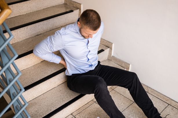 A man sitting on the bottom step of a staircase, holding his lower back in pain.
